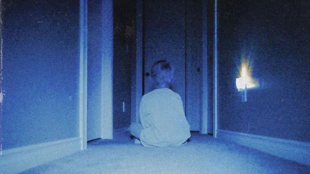 A grainy image of a young boy staring at a slightly-open door, illuminated by a night light. 